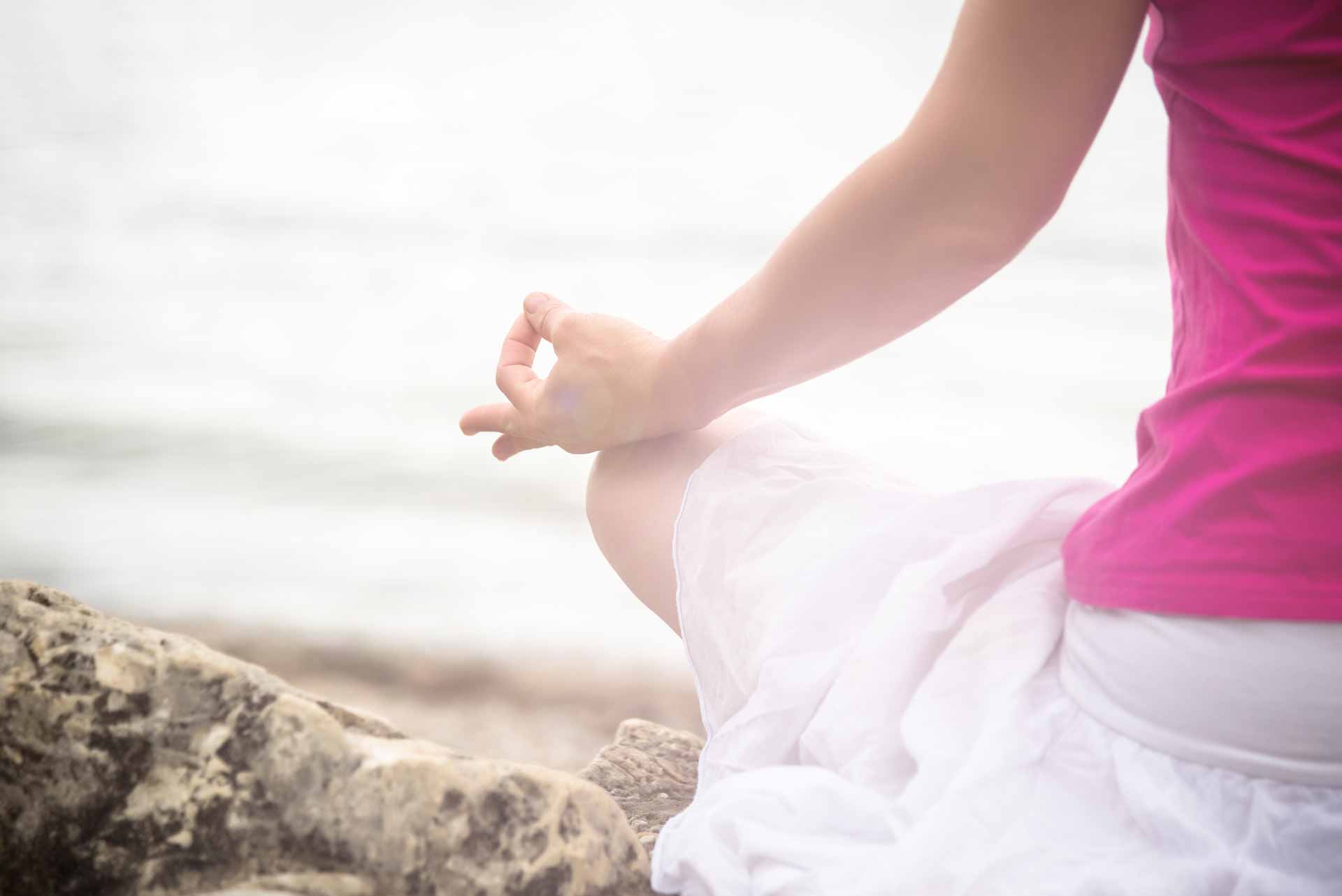 Back view of a woman in a pink shirt and white skirt meditating on the sand by a lake. 
