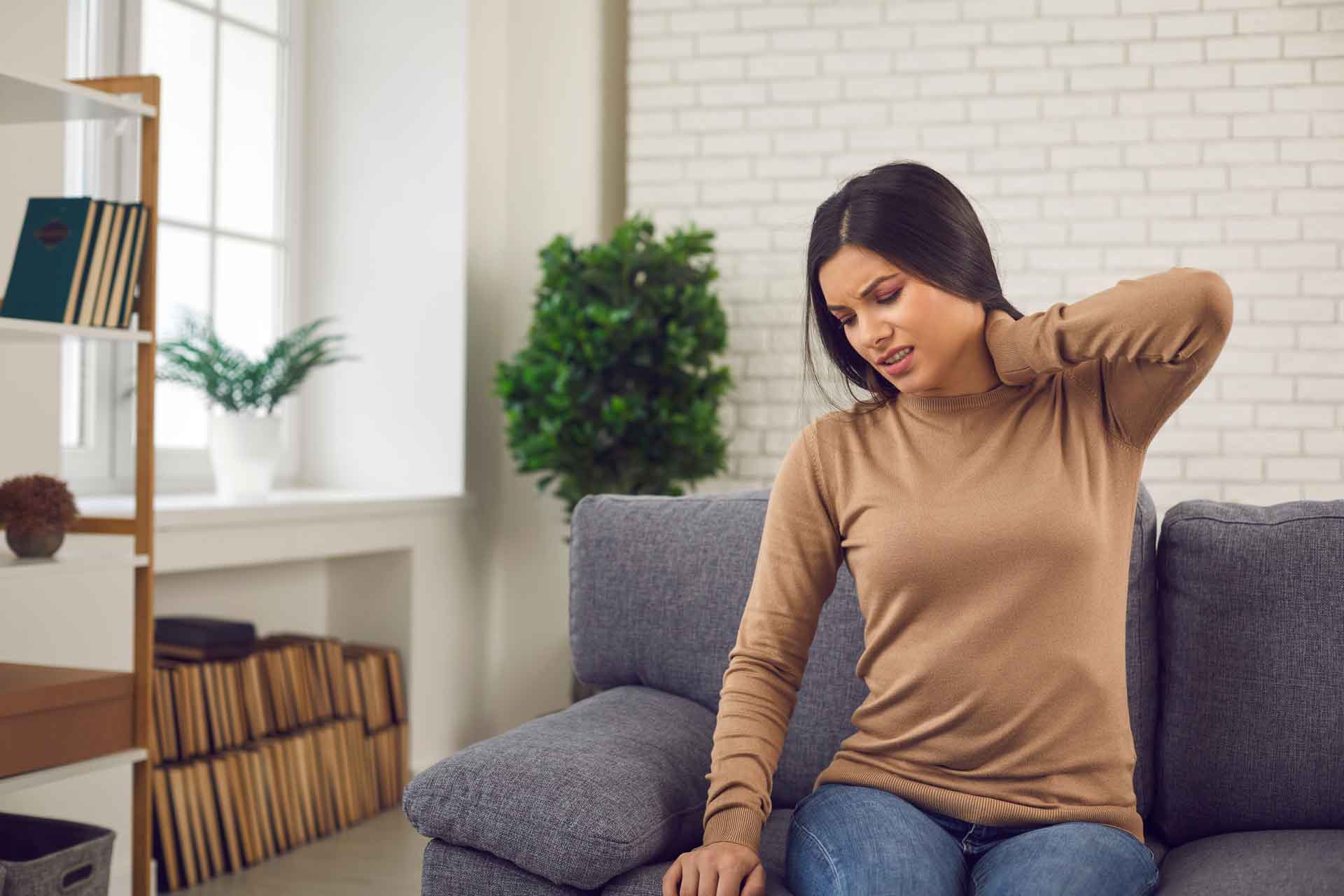 A woman with dark hair and a long-sleeve brown shirt, sitting on a couch and holding her hand to her neck in pain